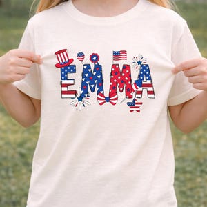 A young girl wears a white personalized patriotic sweatshirt featuring the custom name Emma in large red, white, and blue letters decorated with stars, stripes, fireworks, and a festive patriotic bow. The clean studio background keeps the focus on the custom front design and comfortable fit. This mockup presents a personalized kids sweatshirt perfect for Fourth of July parties, family gatherings, parade outfits, and America 250 celebration apparel.