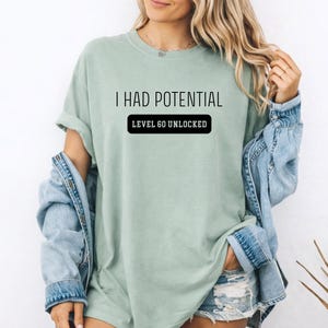May include: A light green t-shirt with the text "I HAD POTENTIAL" above a black rectangle that reads "LEVEL 60 UNLOCKED". The shirt is paired with a denim jacket and shorts.