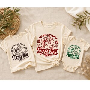 Custom Turkey Trot Squad family shirts for Thanksgiving 2026. Cream colored matching tees and a baby bodysuit feature a retro turkey running graphic, autumn maple leaves, family name, year 2026, and “Turkey Trot Squad” lettering. A festive matching family outfit for Thanksgiving Day races, turkey runs, holiday gatherings, family pictures, and seasonal celebrations.