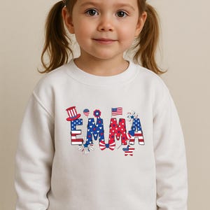 A child points toward the front of a natural cream personalized patriotic T-shirt featuring the custom name Emma in bold red, white, and blue letters with stars, stripes, fireworks, and celebratory American flag-inspired accents. The outdoor background adds a fun summer holiday feel while keeping the design easy to see. This mockup is ideal for showcasing a custom kids Fourth of July shirt made for parades, parties, and America 250 events.