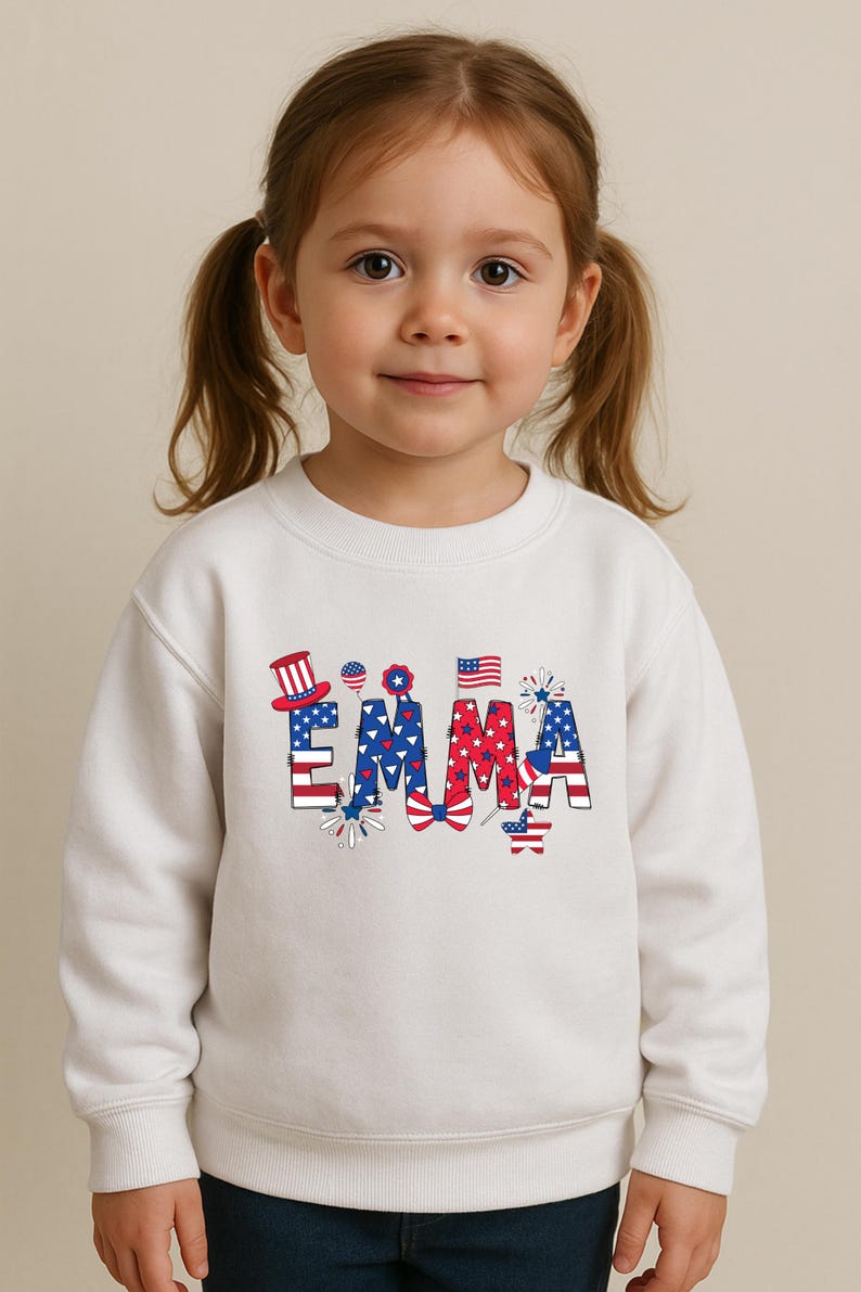 A child points toward the front of a natural cream personalized patriotic T-shirt featuring the custom name Emma in bold red, white, and blue letters with stars, stripes, fireworks, and celebratory American flag-inspired accents. The outdoor background adds a fun summer holiday feel while keeping the design easy to see. This mockup is ideal for showcasing a custom kids Fourth of July shirt made for parades, parties, and America 250 events.