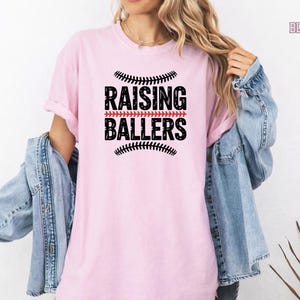 This Raising Ballers Comfort Colors® tee is the perfect game day shirt for sports moms who proudly support their kids on the field. Featuring a classic baseball inspired design and soft garment dyed fabric, this shirt combines comfort and style for baseball season, softball tournaments, and everyday wear.