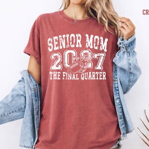 A woman wearing a red t-shirt with the text "SENIOR MOM 2007 THE FINAL QUARTER" printed on it, along with a denim jacket.