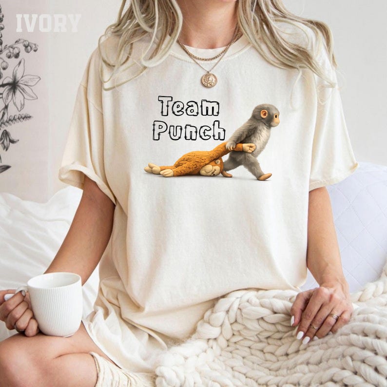 A woman wearing a white t-shirt with a graphic of two cartoon animals, one brown and one gray, and the text "Team Punch" printed on it. She is sitting on a bed with a white blanket.