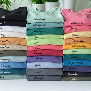 A collection of colorful t-shirts is neatly stacked on a white surface, with each shirt displaying a different color and a handwritten label.