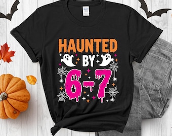 May include: Black t-shirt with the text "HAUNTED BY 6-7" in orange and pink. The design includes white ghosts, spiderwebs, and stars, perfect for Halloween. The shirt is folded with a pair of gray jeans.