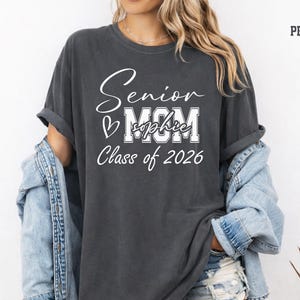 Comfort Colors® senior mom shirt in blue jean color featuring a white Class of 2026 design with bold MOM lettering, script Senior text, heart detail and customizable name across the center. Perfect for proud moms celebrating their graduate’s senior year, graduation events, school photos, senior night, college send off, family gatherings and Class of 2026 memories.