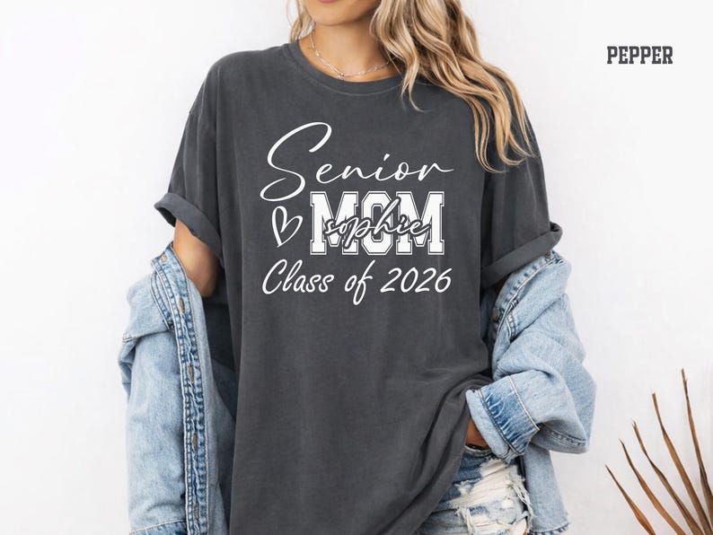 Comfort Colors® senior mom shirt in blue jean color featuring a white Class of 2026 design with bold MOM lettering, script Senior text, heart detail and customizable name across the center. Perfect for proud moms celebrating their graduate’s senior year, graduation events, school photos, senior night, college send off, family gatherings and Class of 2026 memories.