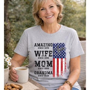 Close-up of a beige t-shirt showcasing a custom “Amazing Wife Mom Grandma Since Year” design with a vintage distressed font. The right side features a bold USA flag print, adding a patriotic touch. Ideal for grandmothers celebrating family milestones, this personalized shirt makes a thoughtful gift for birthdays, Mother’s Day, or family gatherings. The natural fabric color enhances the classic and timeless look of the design.