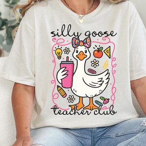A white t-shirt with a cartoon image of a duck holding a drink and surrounded by various school-related items, such as a pencil, apple, and lightbulb.