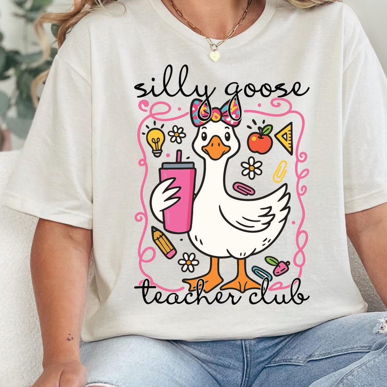 A white t-shirt with a cartoon image of a duck holding a drink and surrounded by various school-related items, such as a pencil, apple, and lightbulb.