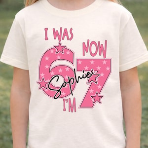 A young girl wearing a white t-shirt with a pink and white design that says "I WAS NOW 67" and "Sophie I'M" printed on it.