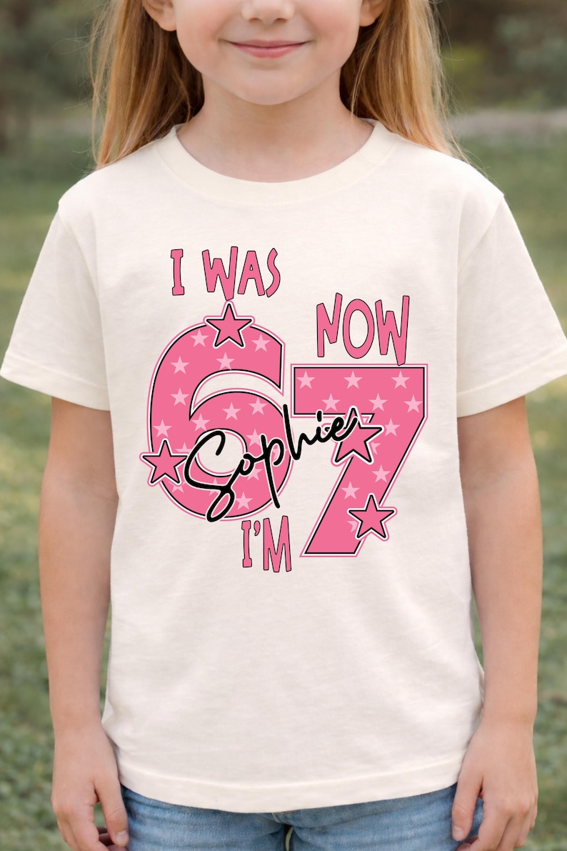 A young girl wearing a white t-shirt with a pink and white design that says "I WAS NOW 67" and "Sophie I'M" printed on it.