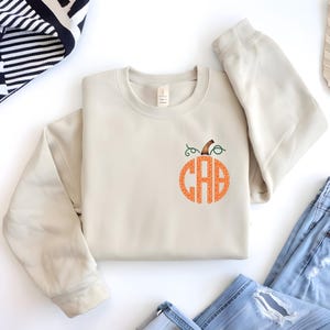 May include: Cream-colored sweatshirt with a pumpkin design. The pumpkin is orange and filled with the letters "CAB". The pumpkin has a green vine and brown stem. The sweatshirt is folded on a white surface with blue jeans and a black and white striped sweater.