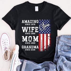 Custom grandma shirt displayed on a cheerful older woman sitting outdoors, wearing a soft neutral-toned tee. The design combines family milestones with a patriotic American flag, creating a meaningful and stylish look. Personalized years make each shirt unique, perfect for gifting to moms or grandmas. This comfortable everyday shirt blends emotional value with trendy distressed typography and classic USA-themed graphics.