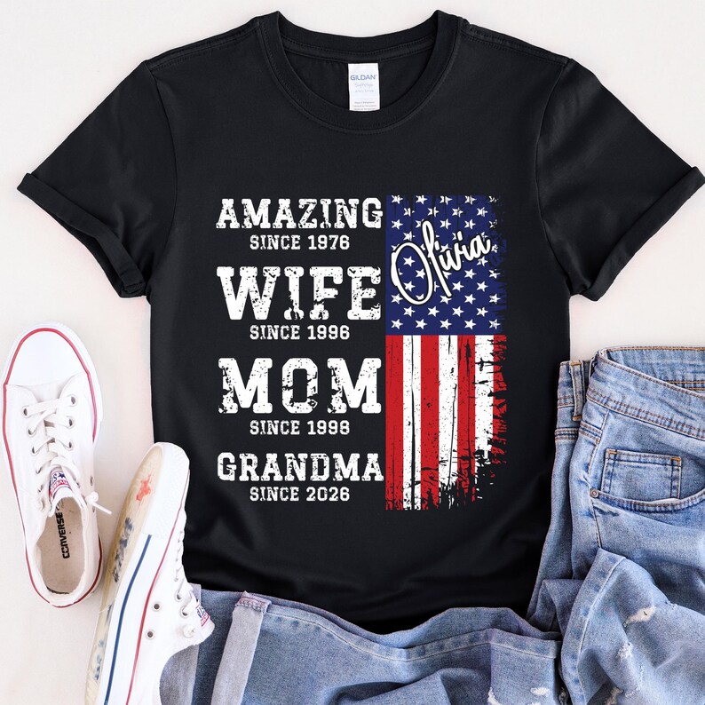 Custom grandma shirt displayed on a cheerful older woman sitting outdoors, wearing a soft neutral-toned tee. The design combines family milestones with a patriotic American flag, creating a meaningful and stylish look. Personalized years make each shirt unique, perfect for gifting to moms or grandmas. This comfortable everyday shirt blends emotional value with trendy distressed typography and classic USA-themed graphics.
