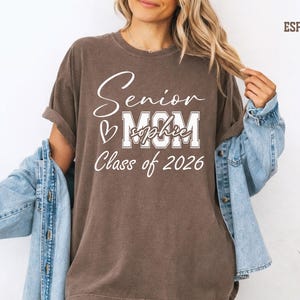 A woman wearing a brown t-shirt with text that says "Senior M.C.M. Class of 2026" and a denim jacket, smiling and posing for the camera.