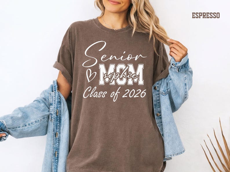 A woman wearing a brown t-shirt with text that says "Senior M.C.M. Class of 2026" and a denim jacket, smiling and posing for the camera.