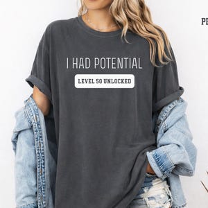 May include: Dark gray t-shirt with the text "I HAD POTENTIAL" above "LEVEL 50 UNLOCKED" in a white rectangle. The shirt is worn with a light blue denim jacket. The word "PEPPER" is printed in the upper right corner.