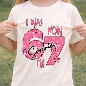 A young girl wearing a white t-shirt with a pink and white design that says "I WAS NOW Sophie I'M" and stars.