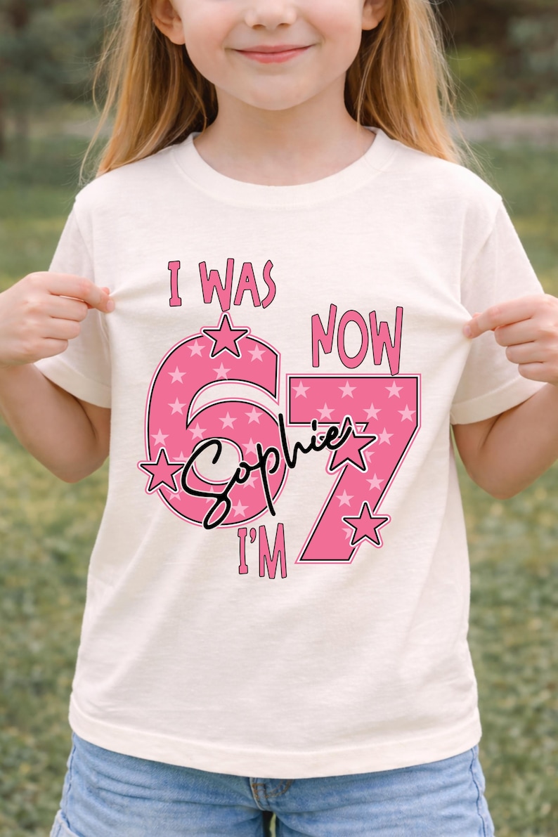 A young girl wearing a white t-shirt with a pink and white design that says "I WAS NOW Sophie I'M" and stars.