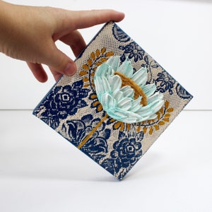 Held in hand for scale view of miniature painting in art deco style with a blue impasto flower on a textured background with golden leaves