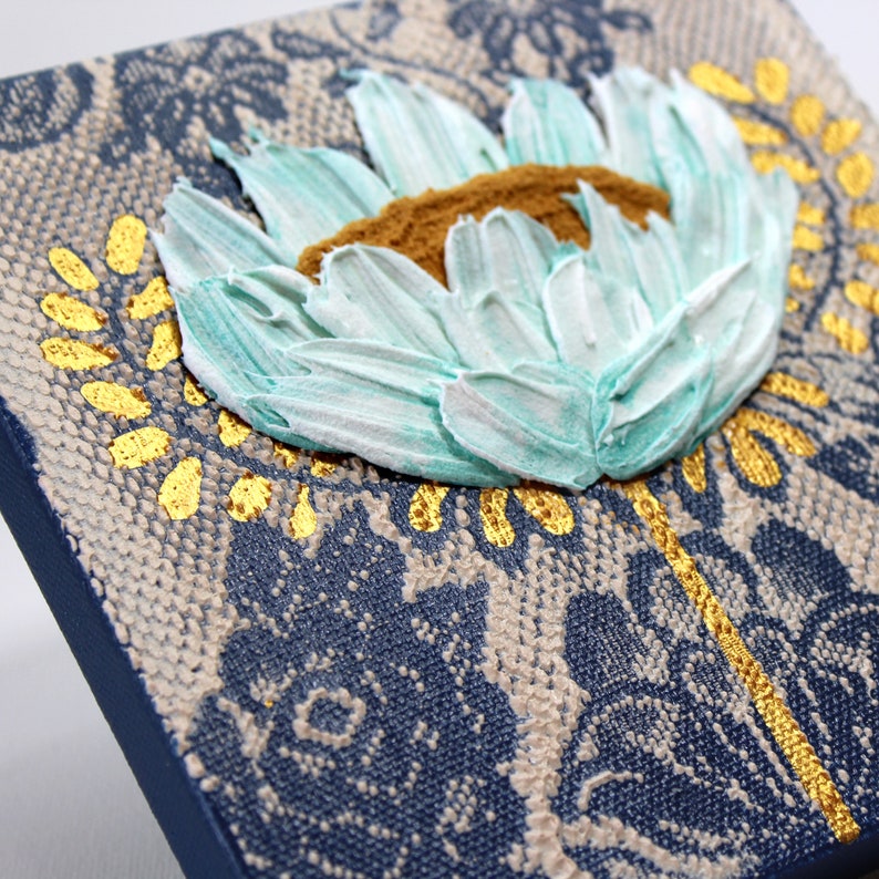 Angled view of miniature painting in art deco style with a blue impasto flower on a textured background with golden leaves