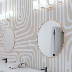 May include: A modern bathroom with two round mirrors with gold frames and a white countertop. The mirrors reflect the room's lighting fixtures. The wallpaper has a beige background with white abstract lines.