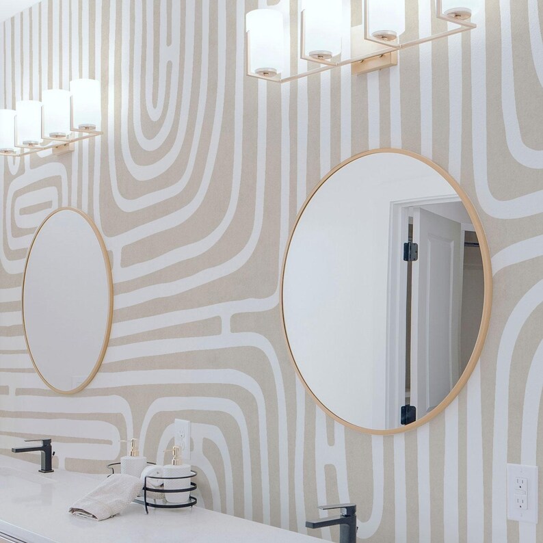 May include: A modern bathroom with two round mirrors with gold frames and a white countertop. The mirrors reflect the room's lighting fixtures. The wallpaper has a beige background with white abstract lines.