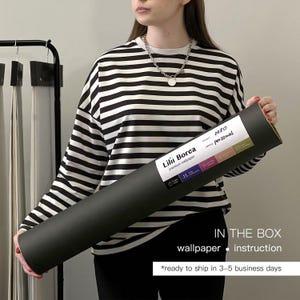 May include: A person holding a rolled-up black wallpaper tube with a white label. The label reads "Lilii Borea premium wallpaper" and includes the text "IN THE BOX wallpaper instruction". The person is wearing a black and white striped long-sleeve shirt.