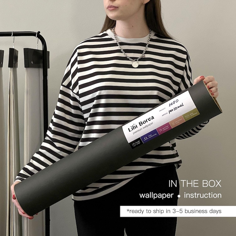 May include: A person holding a rolled-up black wallpaper tube with a white label. The label reads "Lilii Borea premium wallpaper" and includes the text "IN THE BOX wallpaper instruction". The person is wearing a black and white striped long-sleeve shirt.