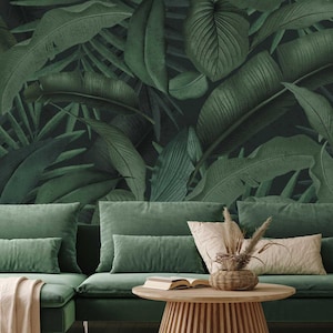 May include: A green velvet sofa with two cushions and a throw, a wooden coffee table with a book and a vase of dried flowers, all in front of a wall with a tropical leaf wallpaper.