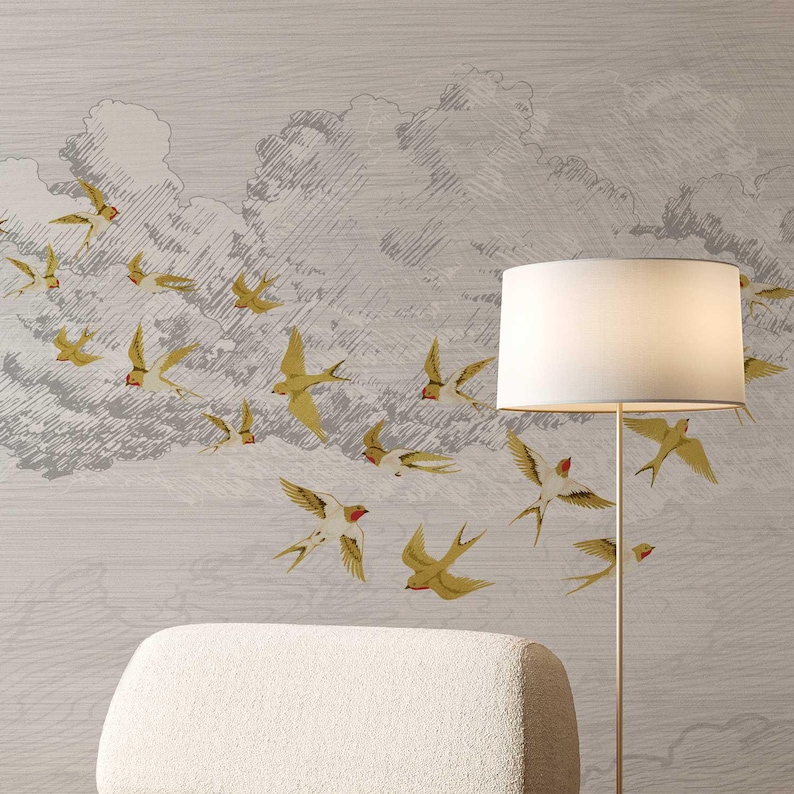 May include: A wall mural featuring a flock of yellow and red-breasted swallows in flight against a gray, cloudy sky. The birds are depicted in a vintage style. A white lamp with a gold base and a cream-colored armchair are in the foreground.