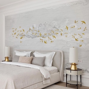 May include: A bedroom scene featuring a bed with a light gray headboard and bedding. The wall behind the bed has a mural of clouds and gold-colored birds in flight. Two bedside lamps with gold bases and white shades flank the bed.