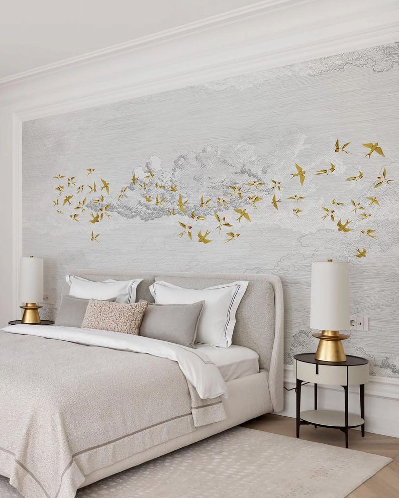 May include: A bedroom scene featuring a bed with a light gray headboard and bedding. The wall behind the bed has a mural of clouds and gold-colored birds in flight. Two bedside lamps with gold bases and white shades flank the bed.