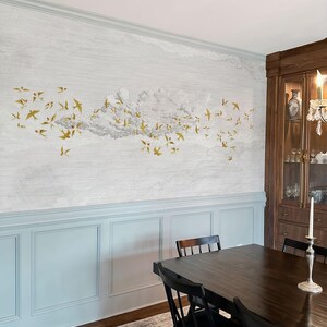 May include: A dining room with a wall mural featuring a flock of gold-colored birds flying across a cloudy sky. The wall is painted light gray with a blue wainscoting. A dark wood dining table and chairs are in the foreground.
