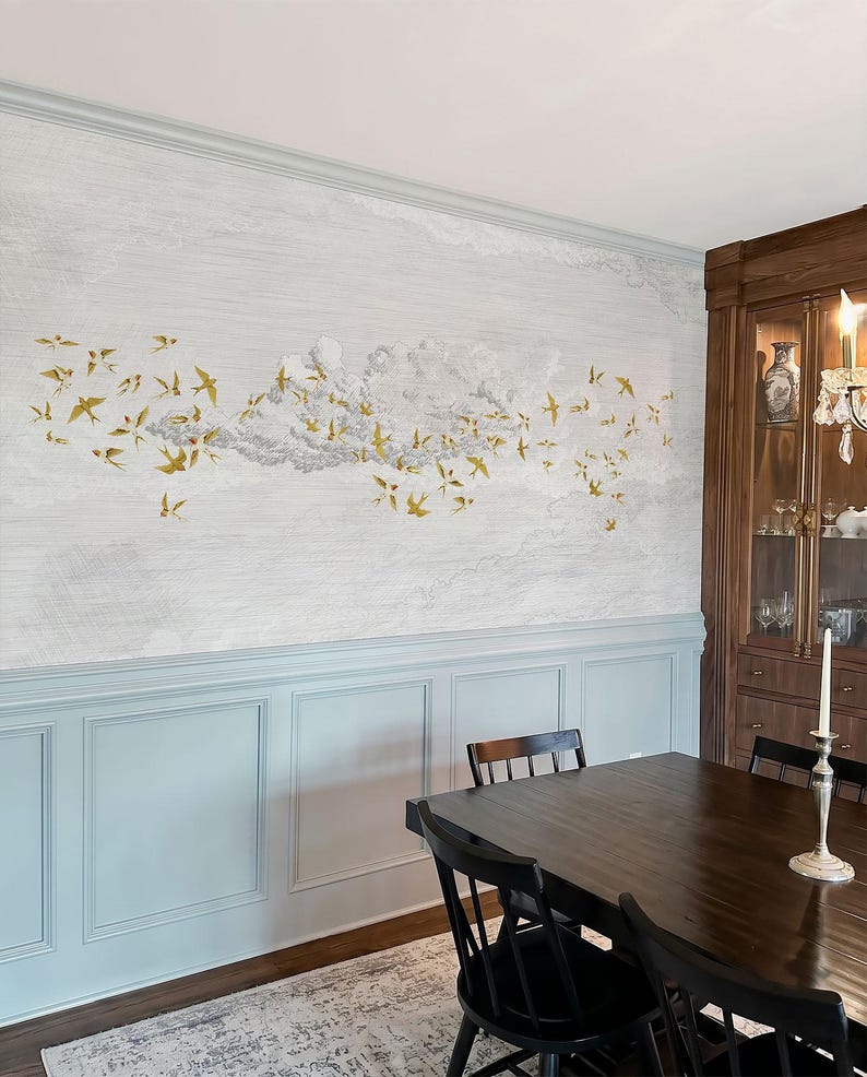 May include: A dining room with a wall mural featuring a flock of gold-colored birds flying across a cloudy sky. The wall is painted light gray with a blue wainscoting. A dark wood dining table and chairs are in the foreground.