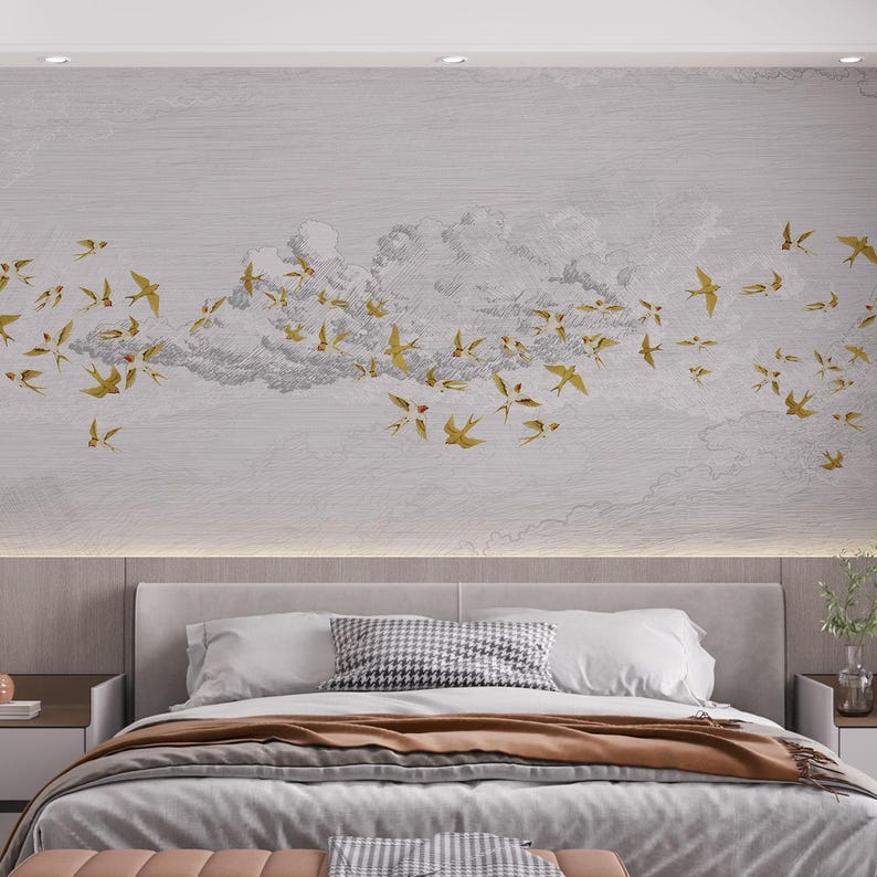 May include: A light gray wall mural featuring a flock of gold-colored birds flying near a cloud formation. The mural is above a bed with white pillows, a brown blanket, and a gray bedspread. A bench is at the foot of the bed.