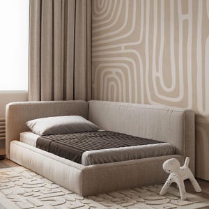 May include: A beige upholstered bed with a brown and gray striped blanket and a white pillow. The bed is in a corner of a room with beige curtains and a patterned wall. A white dog-shaped figurine is on the floor.