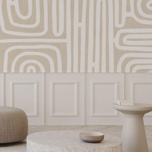 May include: A neutral-toned interior design scene featuring a beige and white abstract patterned wallpaper. Below the wallpaper is a white paneled wall. A round marble coffee table, a small side table, and a beige ottoman are in the foreground.