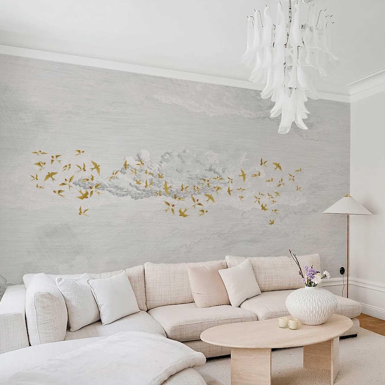 May include: A living room with a light gray wall mural featuring a flock of gold-colored birds flying through a cloud formation. A large, beige sectional sofa with white and blush-colored pillows sits below the mural. A white chandelier hangs from the ceiling.