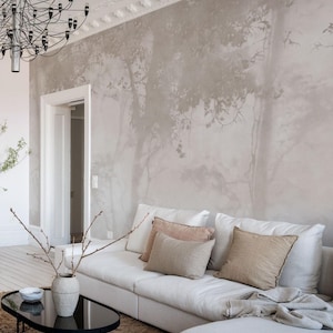 May include: A modern living room with a white sectional sofa, accented with beige and pink pillows. A black oval coffee table sits on a brown rug. The wall features a tree-like mural, and a chandelier hangs from the ceiling.