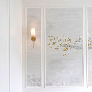 May include: A wall with three framed panels featuring a light gray cloud design and gold bird accents. A brass sconce with a white shade is mounted on the left panel. The room has white trim and a beige carpet.