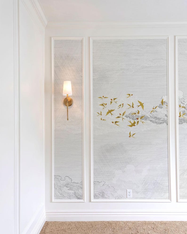 May include: A wall with three framed panels featuring a light gray cloud design and gold bird accents. A brass sconce with a white shade is mounted on the left panel. The room has white trim and a beige carpet.