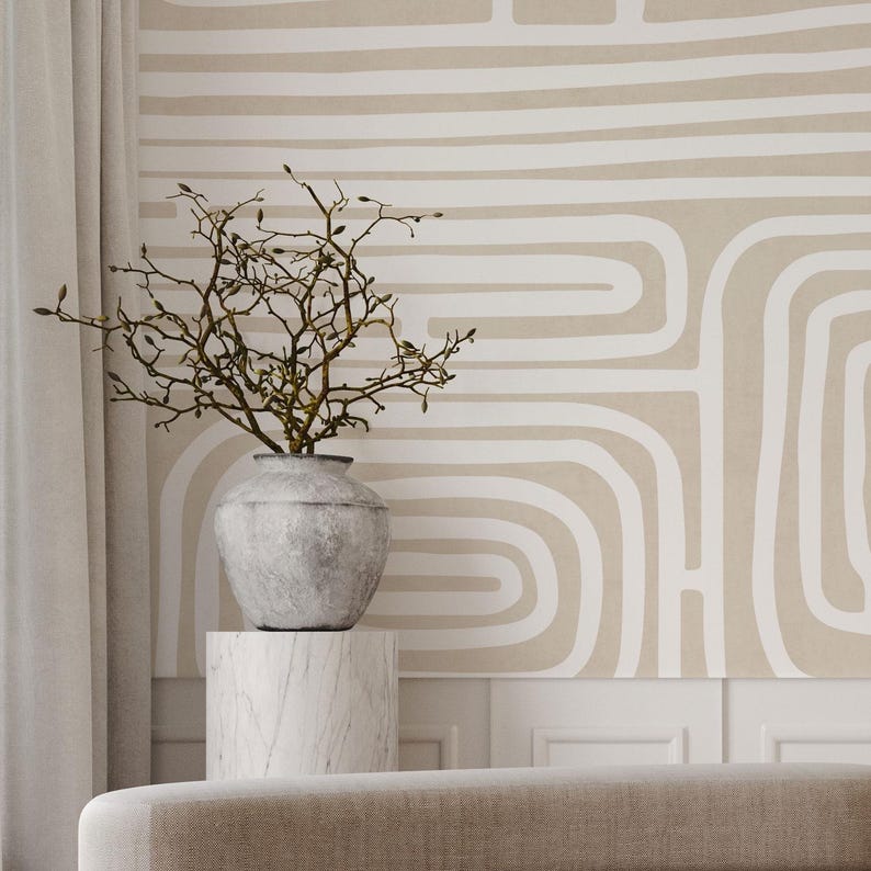 May include: A decorative arrangement featuring a gray vase with branches, set atop a marble pedestal. The background showcases a beige wall with a white abstract pattern. A neutral-toned sofa is in the foreground.