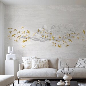 May include: A living room scene featuring a white sofa with fluffy pillows and a black marble coffee table. The wall is adorned with a mural of gold birds flying through a cloud formation. Decorative vases and a sculpture add to the room's aesthetic.