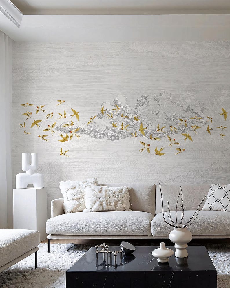 May include: A living room scene featuring a white sofa with fluffy pillows and a black marble coffee table. The wall is adorned with a mural of gold birds flying through a cloud formation. Decorative vases and a sculpture add to the room's aesthetic.