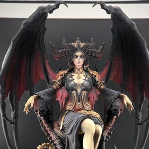 Peut inclure: Une figure féminine à la peau foncée avec des cornes et des ailes est assise sur un trône fait de crânes et d'os. Elle porte une robe noire avec des accents dorés et une cape rouge et noire. Le trône est sur une base circulaire avec une bordure dorée.