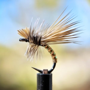 Puede incluir: Primer plano de un señuelo de pesca con mosca marrón y beige con un cuerpo verde y dorado. El señuelo está montado en un soporte de anzuelo negro.