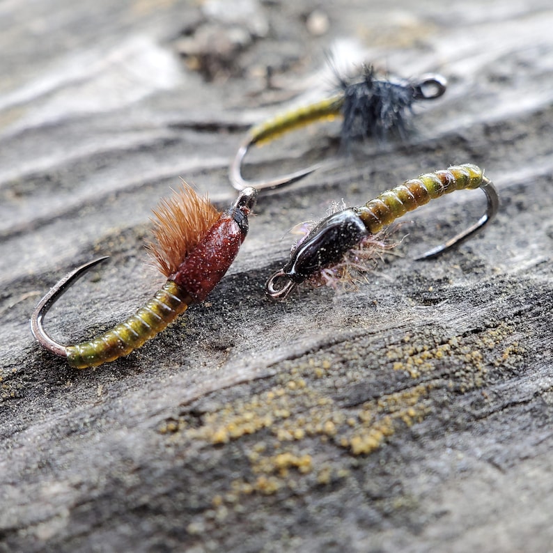May include: Three fly fishing lures with brown, black, and green bodies. The lures are tied with green, brown, and black thread. The lures are on a wooden surface.